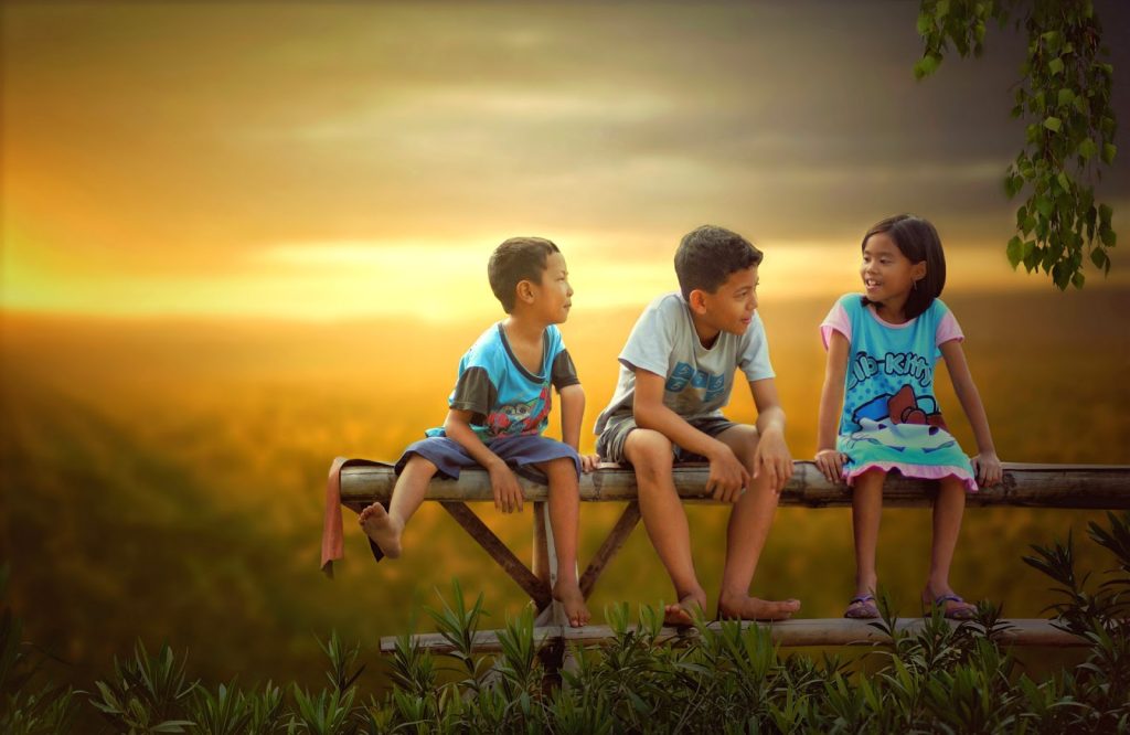 Astra Family Three children developing their social skills as they sit side by side on a wooden bench at sunset.